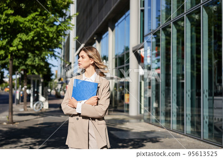 Work and corporate people. Confident businesswoman holding a blue folder with business documents, walking on street Work and corporate people. Confident businesswoman holding a blue folder with business documents, walking on street 95613525
