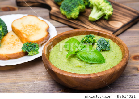 Ready to eat fresh hot broccoli puree soup with slices of broccoli and basil leaves in a wooden plate and slices of toasted white bread on a wooden table. Healthy eating and lifestyle. Ready to eat fresh hot broccoli puree soup with slices of broccoli and basil leaves in a wooden plate and slices of toasted white bread on a wooden table. Healthy eating and lifestyle. 95614021
