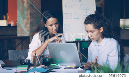 Female coworkers attractive young women are talking and using laptop sitting at desk in shared office. Modern technology, business, teamwork and communication concept. 95614073