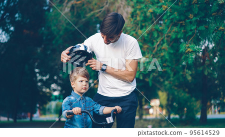 Caring father is putting safety helmet on his little son's head then teaching happy boy to ride bicycle in beautiful park Caring father is putting safety helmet on his little son's head then teaching happy boy to ride bicycle in beautiful park 95614529