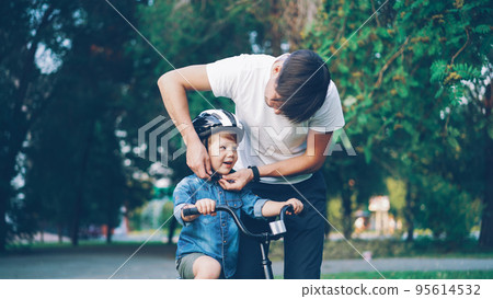 Caring father is putting safety helmet on his little son's head then teaching happy boy to ride bicycle in beautiful park Caring father is putting safety helmet on his little son's head then teaching happy boy to ride bicycle in beautiful park 95614532