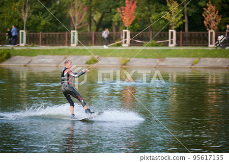 Wakeboarder surfing on lake. Young man surfer having fun wakeboarding in the cable park. Water sport, outdoor activity concept. Wakeboarder surfing on lake. Young man surfer having fun wakeboarding in the cable park. Water sport, outdoor activity concept. 95617155