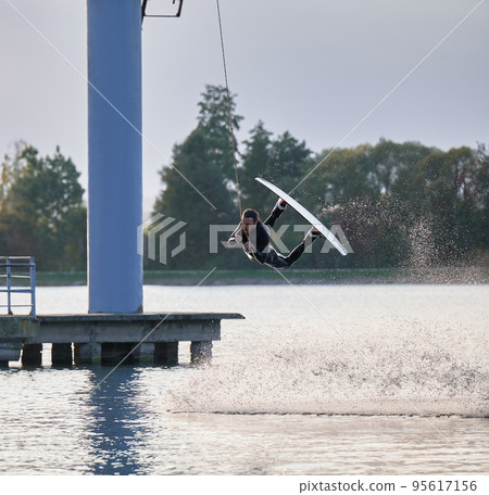 Wakeboarder making tricks while wakeboarding on lake. Young man surfer having fun wakesurfing in the cable park. Water sport, outdoor activity concept. 95617156