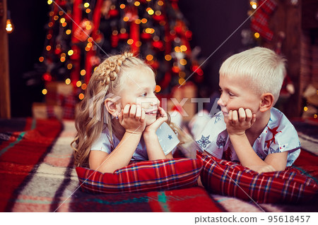 Children in pajamas lying on the floor looking to each other near Christmas tree and fireplace  95618457