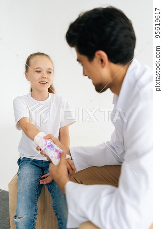 Vertical shot of cheerful little girl with broken hand wrapped in white plaster bandage talking about problem to pediatrician male doctor, during checkup consultation meeting, at medical clinic Vertical shot of cheerful little girl with broken hand wrapped in white plaster bandage talking about problem to pediatrician male doctor, during checkup consultation meeting, at medical clinic 95619617