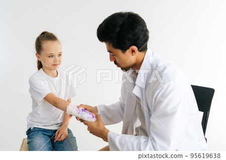 Studio shot of male doctor in uniform consulting little girl patient with broken hand wrapped in plaster bandage at checkup meeting on white isolated background. Concept of insurance and healthcare. Studio shot of male doctor in uniform consulting little girl patient with broken hand wrapped in plaster bandage at checkup meeting on white isolated background. Concept of insurance and healthcare. 95619638
