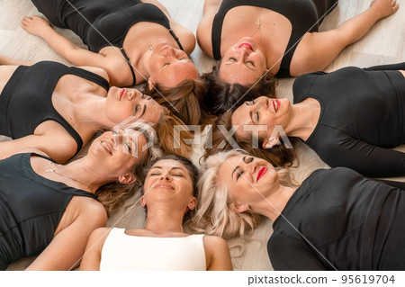 Seven happy women lie on their backs on the floor with their heads pressed together. 95619704