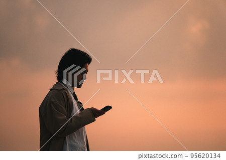 A man looking at a smartphone at the beach in the evening A man looking at a smartphone at the beach in the evening 95620134