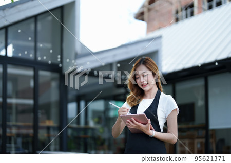 Starting and opening a small business, a young Asian woman showing a smiling face holding a tablet in an apron standing in front of a coffee shop bar counter. Business Owner, Restaurant, Cafe concept. Starting and opening a small business, a young Asian woman showing a smiling face holding a tablet in an apron standing in front of a coffee shop bar counter. Business Owner, Restaurant, Cafe concept. 95621371