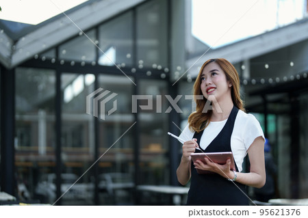 Starting and opening a small business, a young Asian woman showing a smiling face holding a tablet in an apron standing in front of a coffee shop bar counter. Business Owner, Restaurant, Cafe concept. 95621376