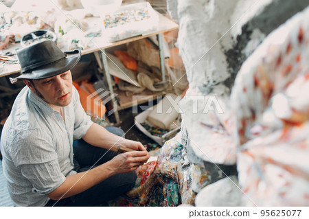 Man mosaics master in hat making smalt glass mosaic panel. Male mosaicist at work 95625077