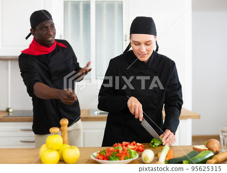 Dissatisfied adult chef controlling work of young female assistant in kitchen Dissatisfied adult chef controlling work of young female assistant in kitchen 95625315