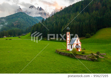 The Church of St. Johann is in a beautiful scenic location against the Geisler peaks covered with clouds in Santa Maddalena village, Val Di Funes, Italy. 95627205