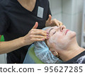 An adult woman of 63 years lies on the procedure of rejuvenation at the beautician with a mask on her face. Cosmetologist's hands massage the lobb. High quality photo 95627285