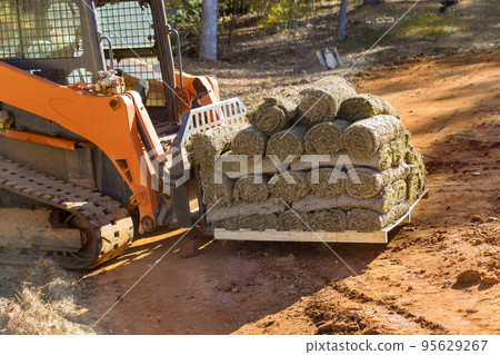 Forklift unloading green grass turf rolls in pallets for landscaping on construction site with used boom truck Forklift unloading green grass turf rolls in pallets for landscaping on construction site with used boom truck 95629267
