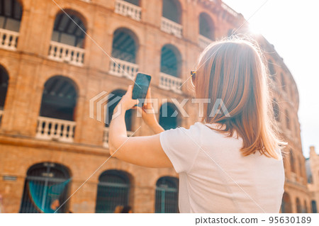 Rear view of blonde young female tourist is exploring new city making photo on smartphone on the old town of Plaza de Toros de Valencia on a bright sunny day Rear view of blonde young female tourist is exploring new city making photo on smartphone on the old town of Plaza de Toros de Valencia on a bright sunny day 95630189