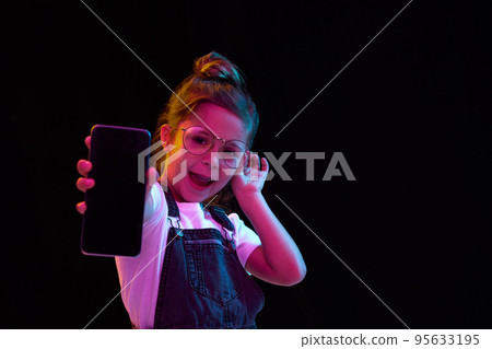 Fashionable nice-looking little girl showing empty phone screen isolated over dark background in neon light. Concept of beauty, kids fashion, children emotions Fashionable nice-looking little girl showing empty phone screen isolated over dark background in neon light. Concept of beauty, kids fashion, children emotions 95633195