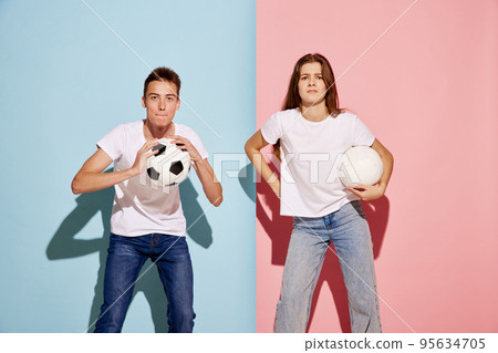 Portrait of young man and woman, sport fans posing with football ball isolated over blue-pink background. Tense moment Portrait of young man and woman, sport fans posing with football ball isolated over blue-pink background. Tense moment 95634705