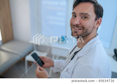 Joyous doctor with the cell phone seated in his office Joyous doctor with the cell phone seated in his office 95634716