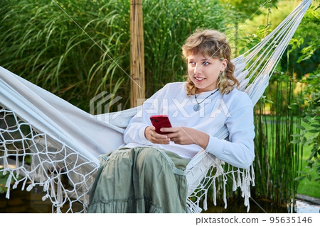 Teenage girl sitting in hammock using smartphone for leisure study communication Teenage girl sitting in hammock using smartphone for leisure study communication 95635146