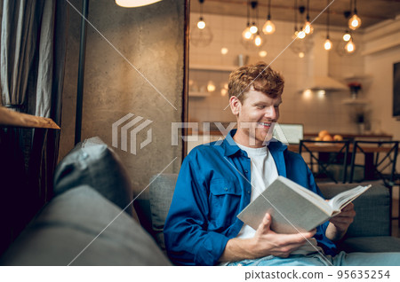 Ginger young man sitting at home at the window with a book Ginger young man sitting at home at the window with a book 95635254