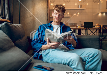 Ginger young man sitting at home at the window with a book Ginger young man sitting at home at the window with a book 95635255