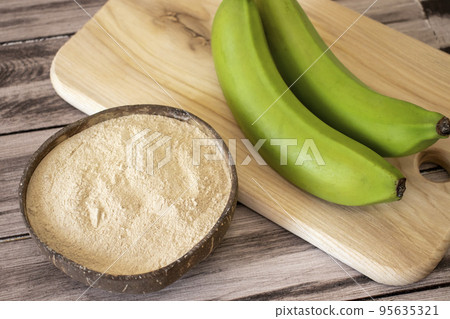 Banana brown flour in a natural bowl on a wooden surface with fresh green bananas 95635321