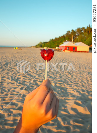 Red candy in the shape of a heart in a hand on sandy beach background. Sunset warm light. Vertical shot. 95637281