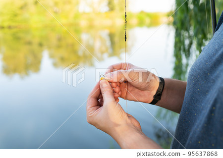 The concept of bait for fish. Close-up of a fisherman's male hand stringing bait on a fishing rod against a blue lake. The concept of bait for fish. Close-up of a fisherman's male hand stringing bait on a fishing rod against a blue lake. 95637868
