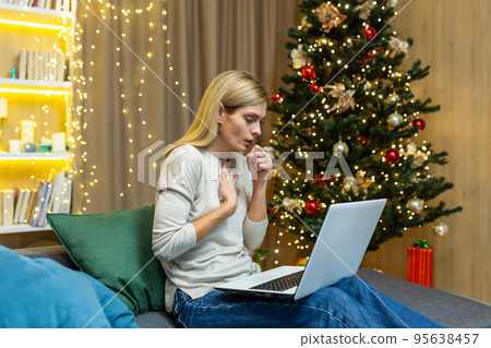 Woman sick on Christmas working with laptop on New Year holidays coughing sitting on sofa at home in living room near Christmas tree. 95638457