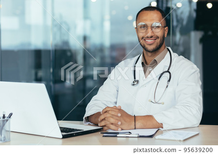 Portrait of young hispanic male pediatrician doctor. Sitting in the office at a table with documents and a laptop, wearing glasses and a stethoscope in a white coat, looking at the camera, smiling. Portrait of young hispanic male pediatrician doctor. Sitting in the office at a table with documents and a laptop, wearing glasses and a stethoscope in a white coat, looking at the camera, smiling. 95639200