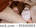 A young girl is fast asleep lying on her stomach with her face in a pillow with her hair down in bed at home.  95640869