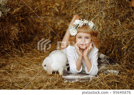 A cute and beautiful little girl is sitting in the hayloft with a rabbit 95640876