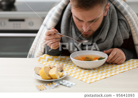 Sick young man eating tasty soup to cure flu at table in kitchen Sick young man eating tasty soup to cure flu at table in kitchen 95641093