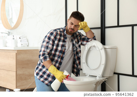 Young man feeling disgust while cleaning toilet bowl in bathroom Young man feeling disgust while cleaning toilet bowl in bathroom 95641362