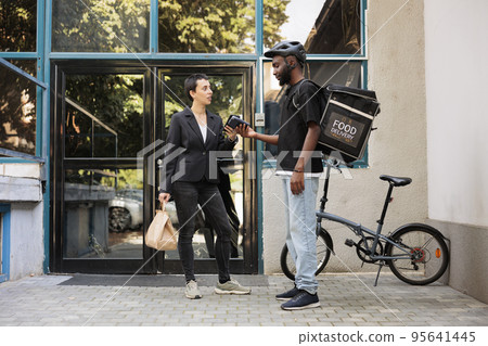 Woman paying for office food delivery service with smartphone, contactless pos terminal, mobile wallet payment. Courier delivering restaurant takeaway meal employee in front of company building Woman paying for office food delivery service with smartphone, contactless pos terminal, mobile wallet payment. Courier delivering restaurant takeaway meal employee in front of company building 95641445