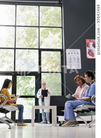 Health specialist discussing with senior woman in hospital waiting room, filling report papers explaining healthcare treatment. Nurse doing checkup examination with asian patient in lobby. 95641494