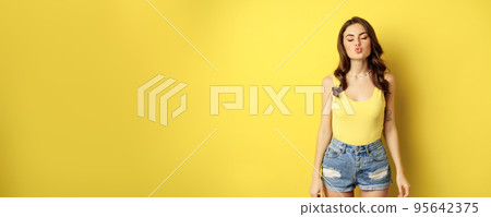 Beautiful, romantic brunette woman kissing, pucker lips, sending air kiss, mwah gesture, standing coquettish against yellow summer background Beautiful, romantic brunette woman kissing, pucker lips, sending air kiss, mwah gesture, standing coquettish against yellow summer background 95642375