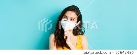 Covid-19, coronavirus and social distancing. Close up portrait of thoughtful attractive woman in respirator mask, looking at upper right corner pensive, blue background Covid-19, coronavirus and social distancing. Close up portrait of thoughtful attractive woman in respirator mask, looking at upper right corner pensive, blue background 95642486
