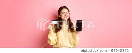 Technology and online shopping. Portrait of beautiful lady showing mobile phone screen and plastic credit card, smiling pleased, recommend app, standing over pink background 95642561