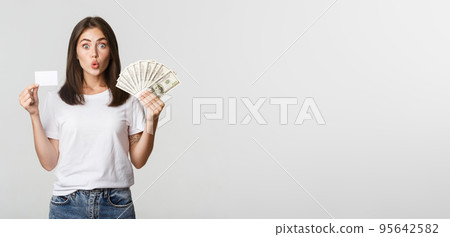 Amused brunette girl looking excited, holding money and credit card, standing white background 95642582