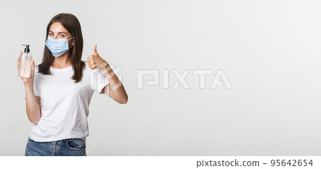 Covid-19, health and social distancing concept. Portrait of satisfied smiling young girl in medical mask, showing thumbs-up and hand sanitizer 95642654
