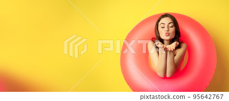 Portrait of beautiful female model posing with pink swimming beach ring, sending air kiss, mwah flirty, standing over yellow background Portrait of beautiful female model posing with pink swimming beach ring, sending air kiss, mwah flirty, standing over yellow background 95642767
