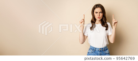 Frustrated young woman pointing fingers up, frowning displeased, standing in white t-shirt over beige background Frustrated young woman pointing fingers up, frowning displeased, standing in white t-shirt over beige background 95642769