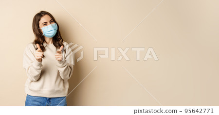 Hey you congrats. Smiling beautiful woman in medical face mask, pointing fingers at camera, standing over beige studio background Hey you congrats. Smiling beautiful woman in medical face mask, pointing fingers at camera, standing over beige studio background 95642771