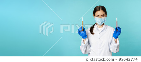 Image of young woman doctor, lab worker doing research, holding test tubes, wearing medical face mask and rubber gloves, blue background Image of young woman doctor, lab worker doing research, holding test tubes, wearing medical face mask and rubber gloves, blue background 95642774