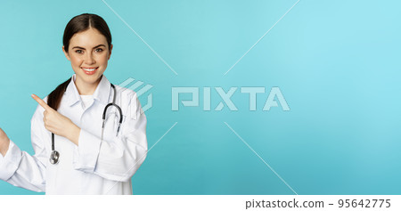 Portrait of smiling medical worker, girl doctor in white coat with stethoscope, pointing fingers left, showing medical clinic advertisement, torquoise background Portrait of smiling medical worker, girl doctor in white coat with stethoscope, pointing fingers left, showing medical clinic advertisement, torquoise background 95642775