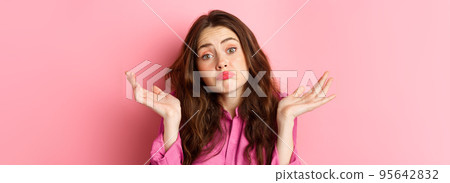 Close up portrait of clueless woman looking coy and unaware, shrugging shoulders, know nothing, cant understand, standing over pink background 95642832
