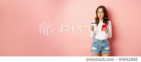 Online shopping. Young woman order online, making purchase in app, holding smartphone and credit card, standing against pink background 95642866
