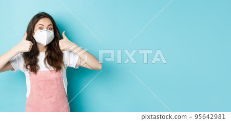Covid, health and pandemic concept. Cheerful young woman in respirator showing thumbs up in approval, recommend something, standing against blue background 95642981